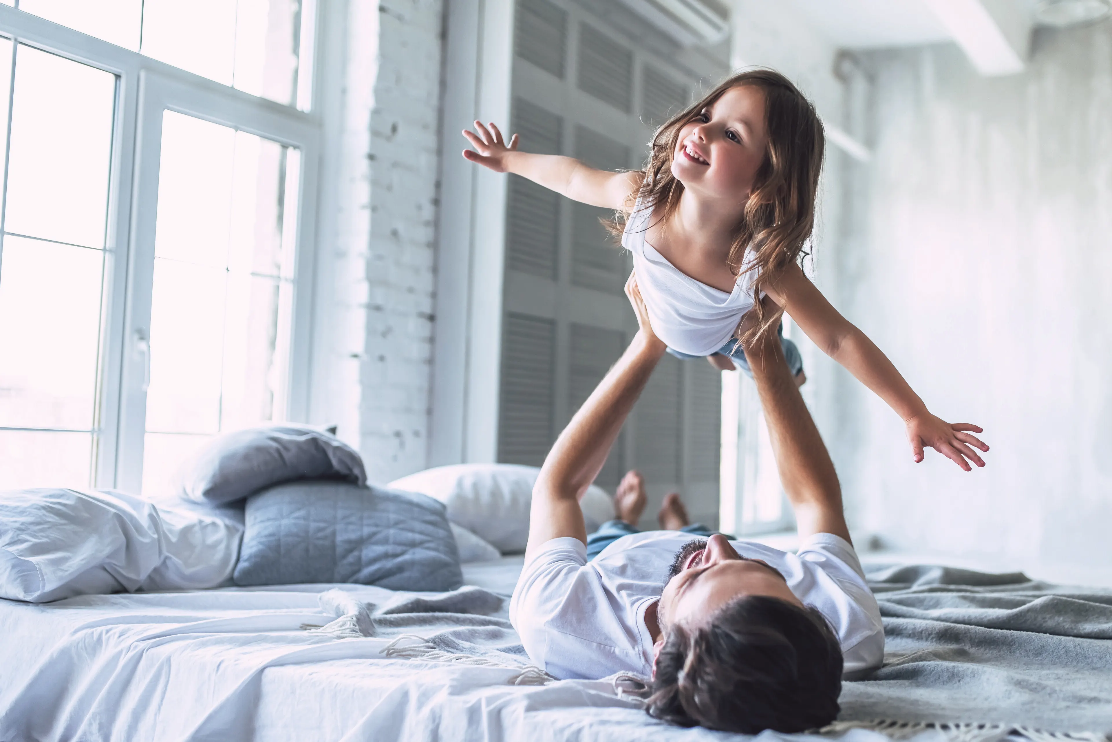 Un père, allongé sur le lit, fait voler sa fille en riant